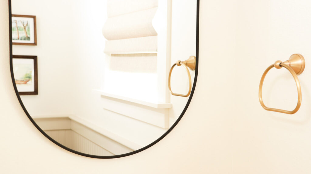 A powder room with an antique brass hand towel ring in front of a thin black, rounded frame mirror showing the reflection of two framed watercolor paintings